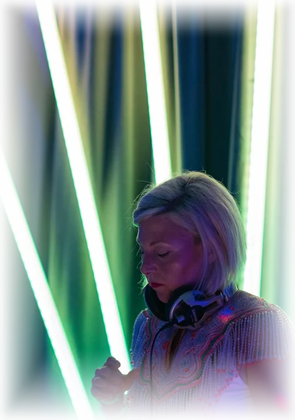 Daniela at the decks with headphones around her neck, lit by tall green and white light tubes